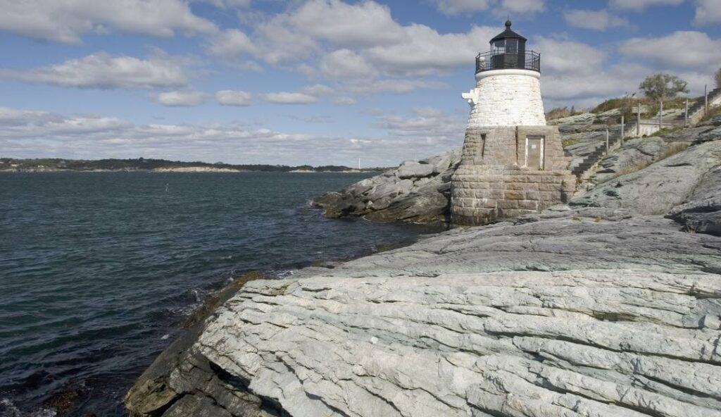 A lighthouse stands on a rocky shoreline overlooking a body of water, with a coastline and cloudy sky visible in the background, much like the dependable beacon that Managed IT Services Rhode Island provides to businesses navigating through their tech challenges.