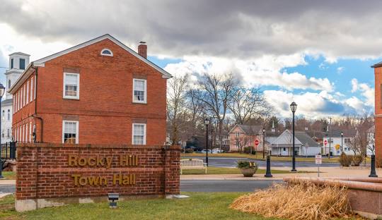 A red-brick building with a sign in front that reads "Rocky Hill Town Hall." The sky is partly cloudy, and other buildings and trees are visible in the background, symbolizing the community where managed IT services in Rocky Hill, CT thrive.