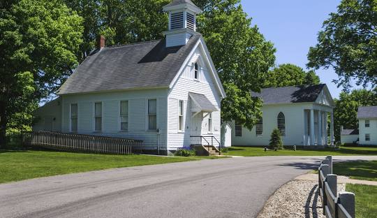 A small white church with a steeple stands near a paved road. Trees and another white building are visible in the background under a clear sky, representing the tranquil charm of Salem, CT.