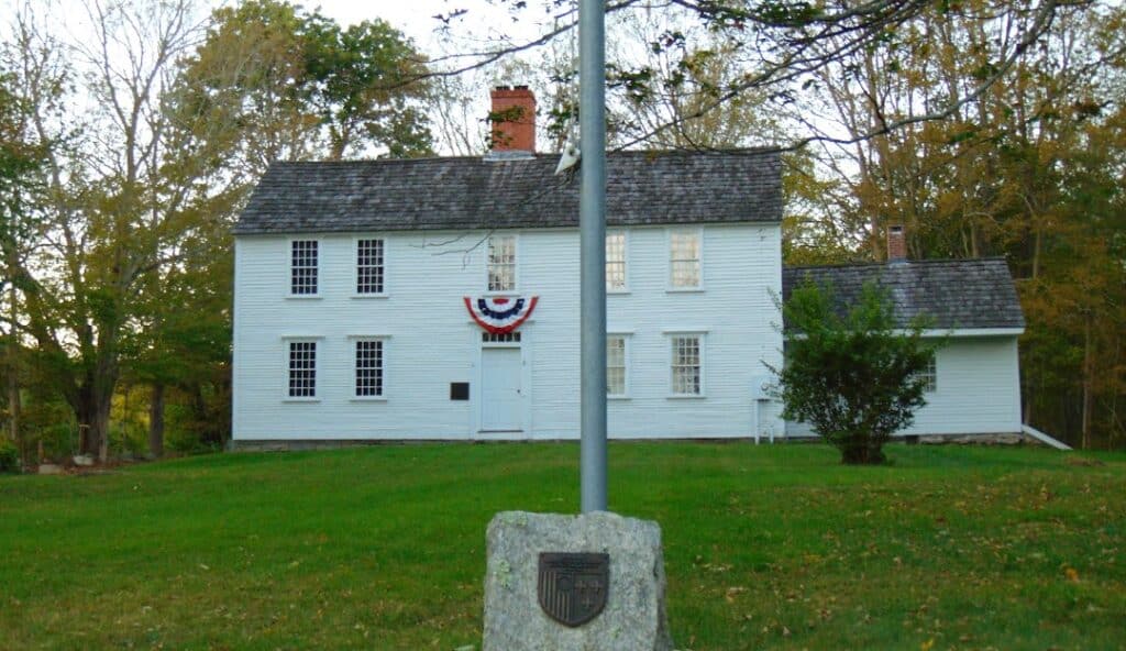 A white colonial-style house with a gray shingled roof, adorned with red, white, and blue bunting stands proudly in Scotland CT. A flagpole with a stone base graces the lawn in front, surrounded by trees. The scene embodies classic charm and meticulous maintenance akin to managed IT services.