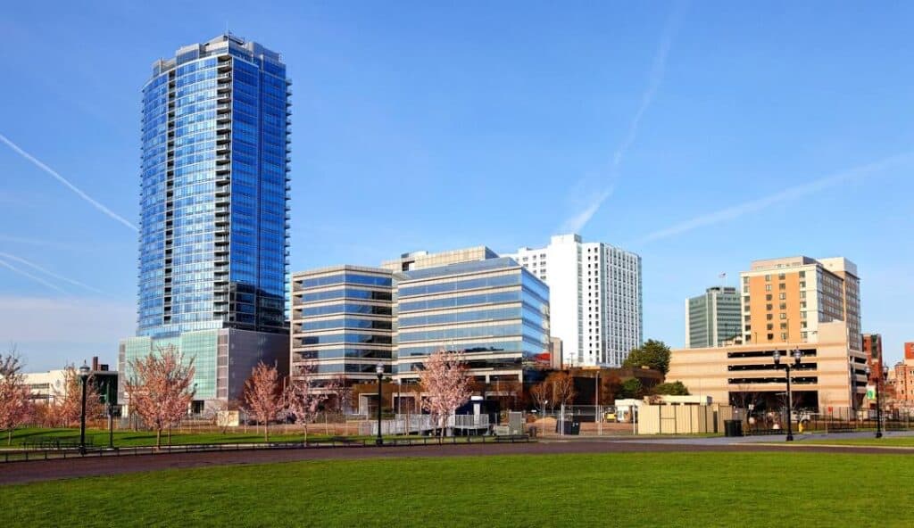 A modern cityscape featuring a tall glass skyscraper and several mid-rise office buildings, viewed from a green park under a clear blue sky, showcases the vibrant hub of managed IT services in Stamford, CT.