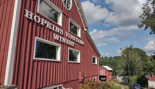 A two-story red barn with white lettering reading "Hopkins Vineyard Winery" set against a backdrop of trees and a clear sky with some clouds, offering picturesque views in Washington CT. Another smaller red barn is visible further in the background.