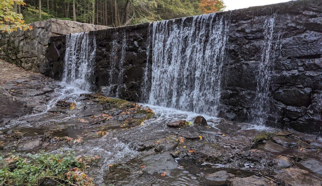 A stone wall with multiple cascading waterfalls flows over rocks and vegetation into a stream with trees in the background, resembling the tranquility of managed IT services Winchester CT provides.