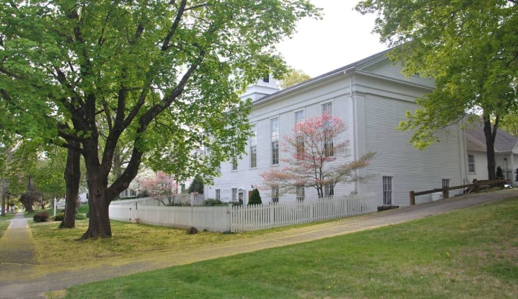 White wooden building with a picket fence, surrounded by trees and a grassy area, viewed from an angle on a slightly inclined path in charming Woodbury CT.