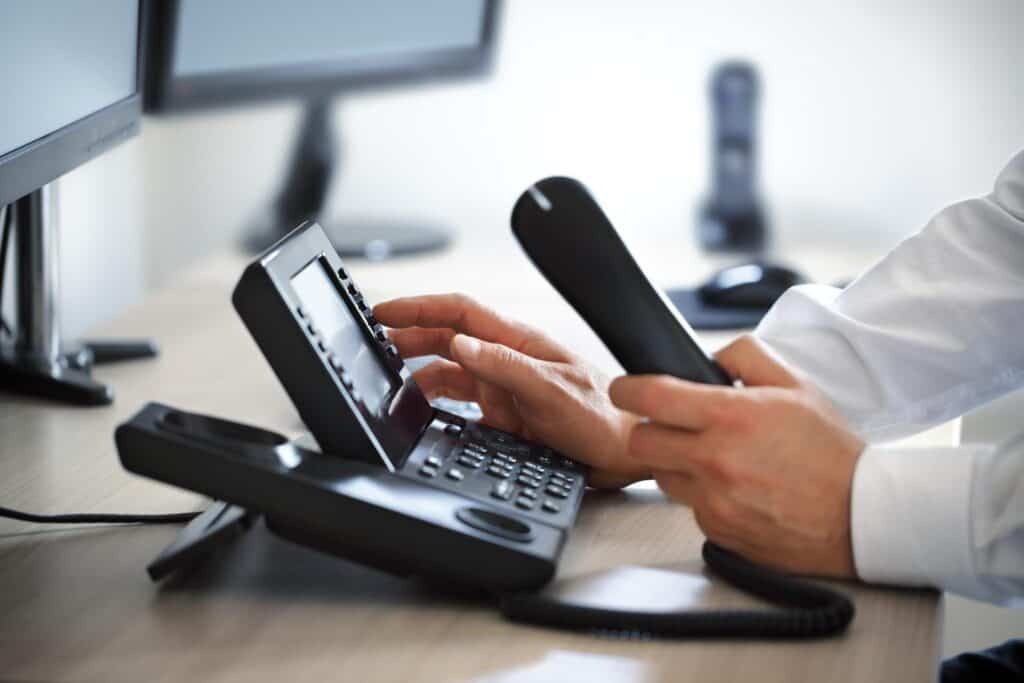 Person dialing a number on a desk phone while holding the receiver, with two computer monitors in the background.