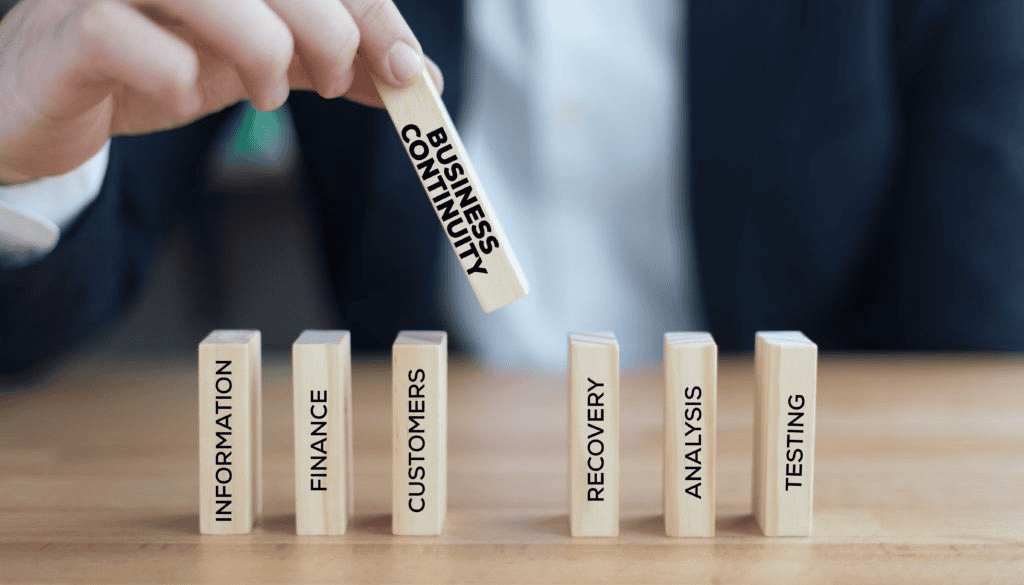 Person holding a wooden block labeled "Business Continuity" above other blocks labeled "Information," "Finance," "Customers," "Recovery," "Analysis," and "Testing.