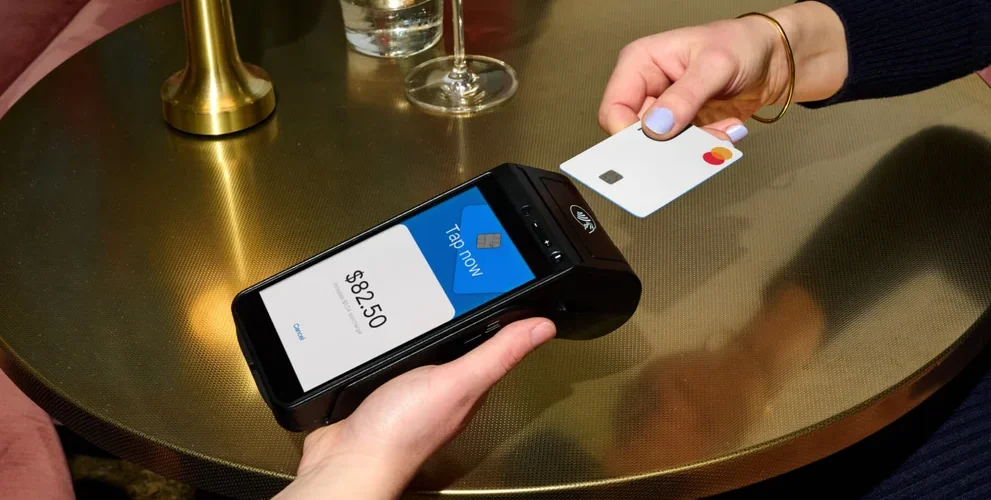 A person holds a credit card near a payment terminal displaying a charge of $82.50, mindful of upcoming 2024 cybersecurity compliance requirements.