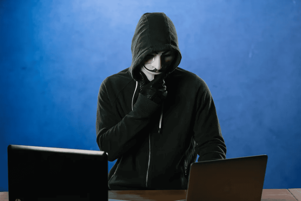 A person in a hoodie and mask sits at a desk between two laptops against a blue background, possibly orchestrating an RIBridges ransomware attack.