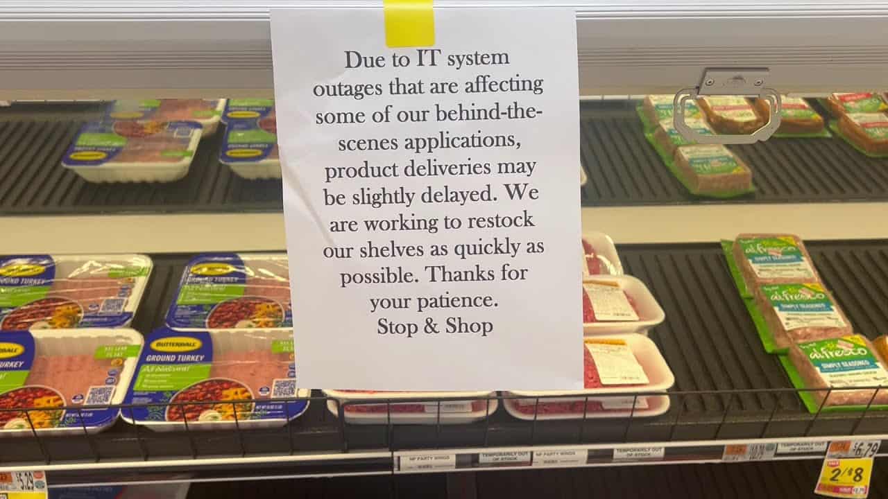 A sign in the grocery store informs customers that IT system outages, related to a Sophos XGS Firewall Upgrade, are causing delays in product deliveries. The store assures that restocking is underway.