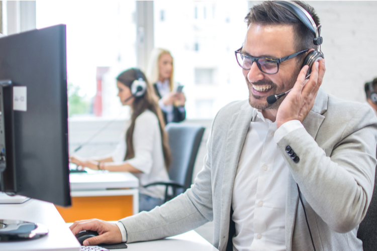 Man in a headset smiling at his computer while providing Managed IT and Network Services in a modern office, with other people wearing headsets in the background.