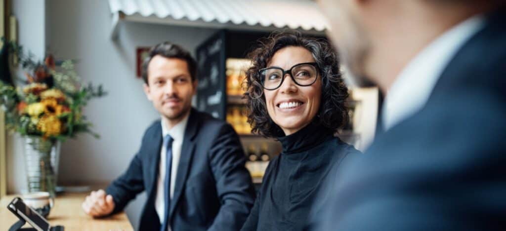 Three Triton Technologies partners in business attire sit at a table in a modern office space, engaged in conversation. One woman is smiling and wearing glasses.