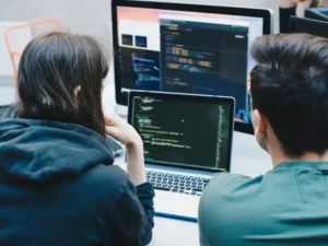 Two people sit at a desk, collaborating on a laptop displaying code, with two large monitors in the background—one showing programming interfaces and the other featuring a project timeline.