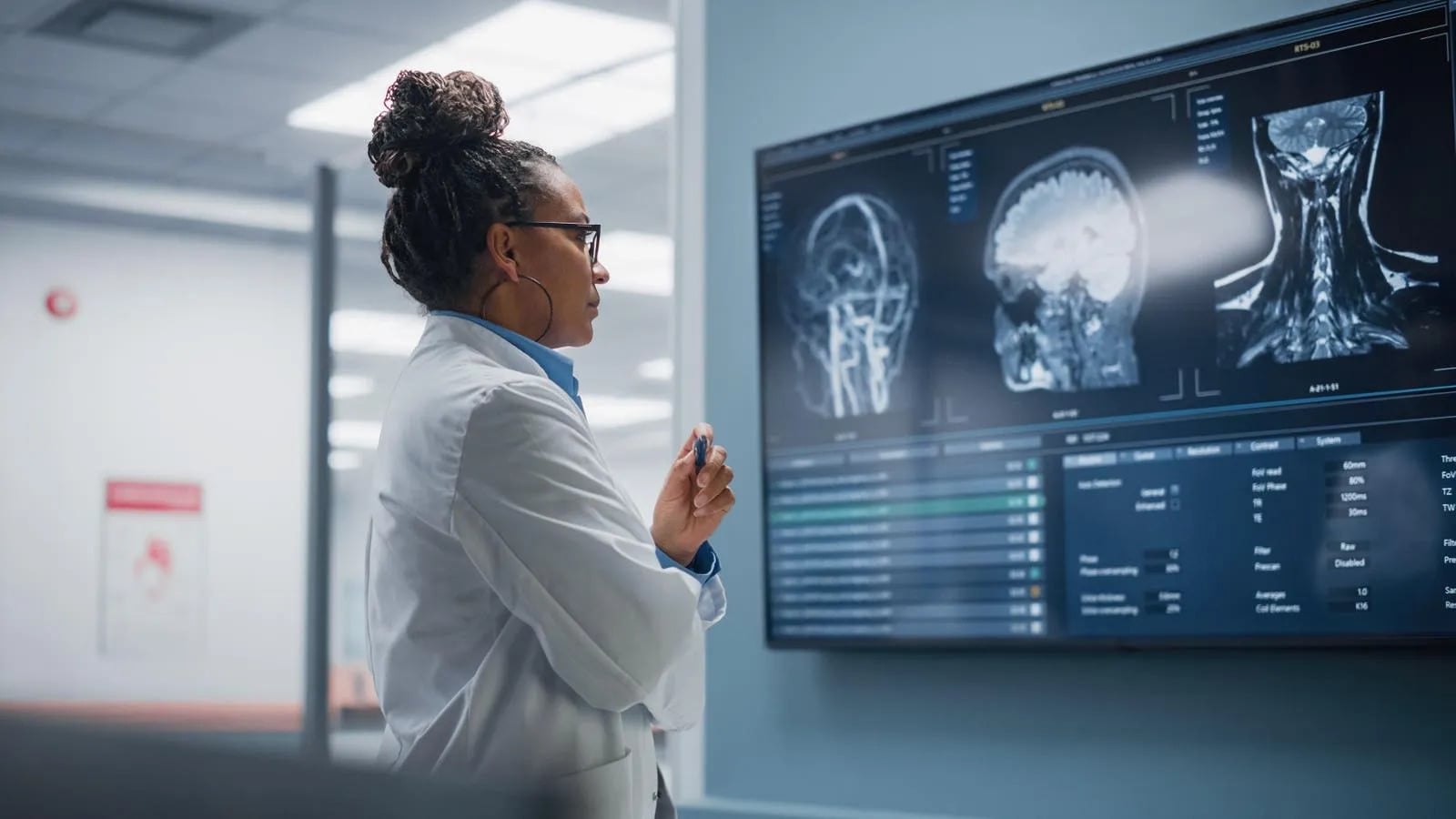 A doctor in a white coat examines brain and neck MRI scans displayed on a large screen in a medical office.