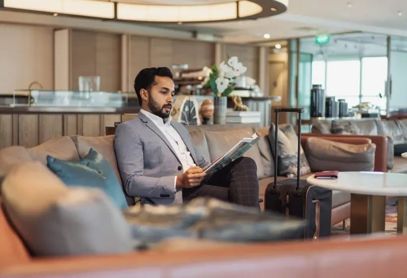 A man in a suit sits on a sofa in an airport lounge, reading a magazine with his suitcase beside him.