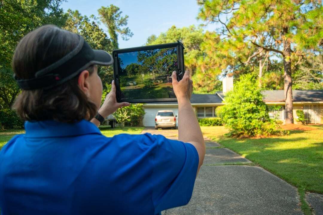 A person in a blue shirt holds up a tablet to take a photo of a house and driveway surrounded by trees and greenery.