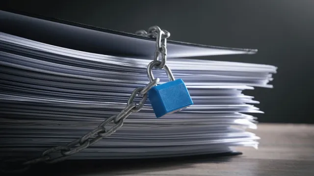 A stack of papers secured with a metal chain and blue padlock, symbolizing restricted access or confidentiality and highlighting the importance of IRS Publication 4557 safeguarding taxpayer data.