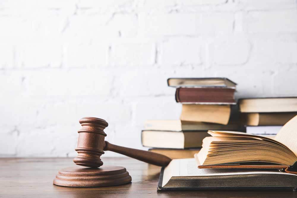 A wooden gavel rests on a block beside an open book and several stacked books on a wooden table against a white brick wall.
