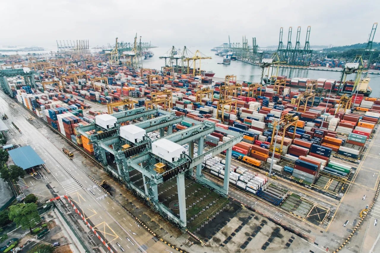 Aerial view of a busy container port with numerous shipping containers, cranes, and docked cargo ships by the waterfront on a cloudy day.