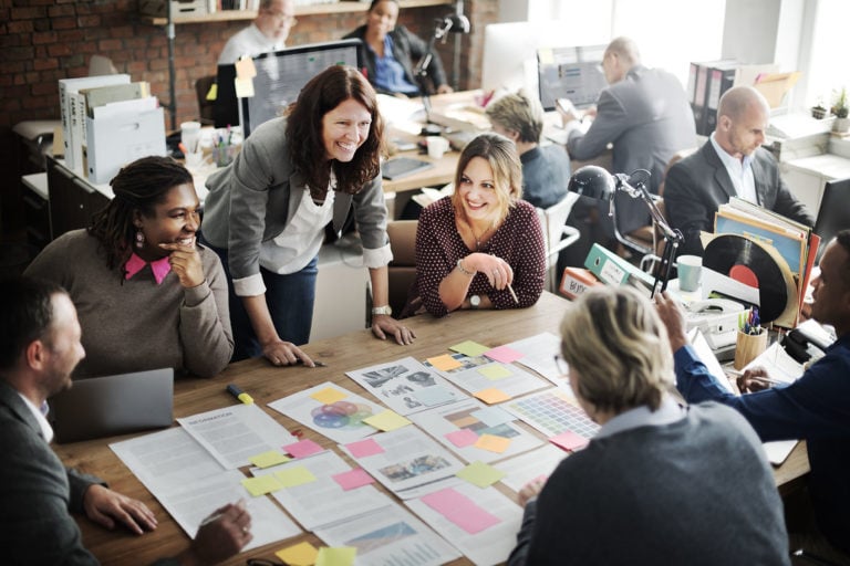 A group of people in business attire sit and stand around a table covered with documents, charts, and sticky notes, engaged in a team meeting in an office.