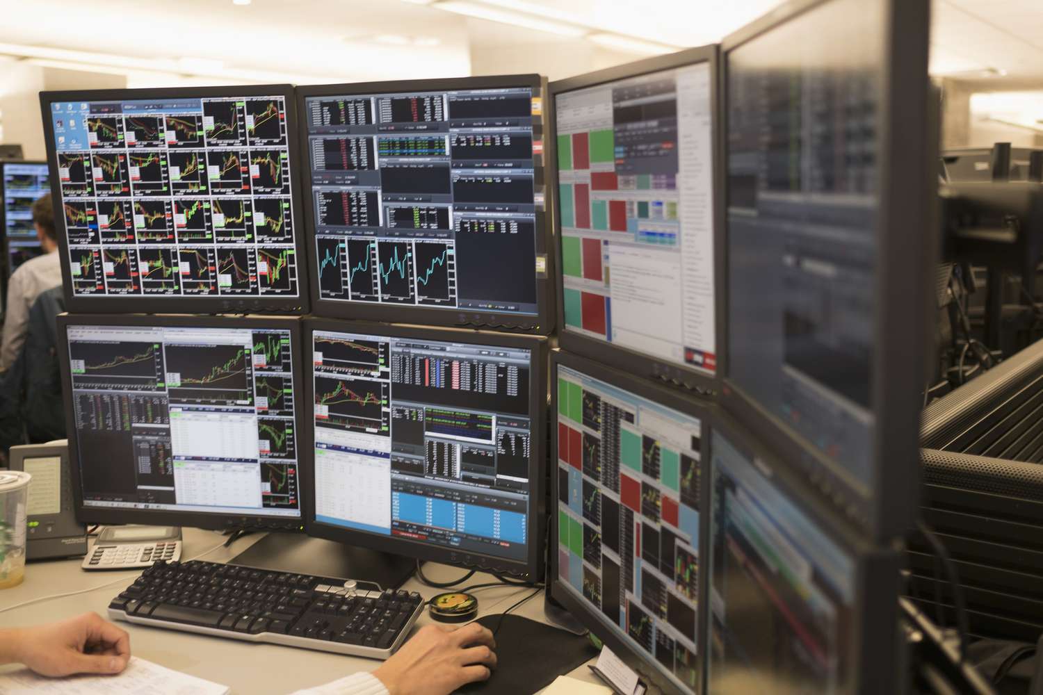 Multiple computer monitors display various financial charts, graphs, and data in a trading office. A person’s hand is on a mouse at the desk.
