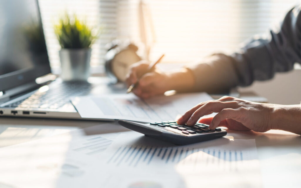 Person using a calculator and writing on financial documents at a desk with a laptop, plant, and alarm clock in the background, demonstrating steps from IRS Publication 4557 safeguarding taxpayer data.
