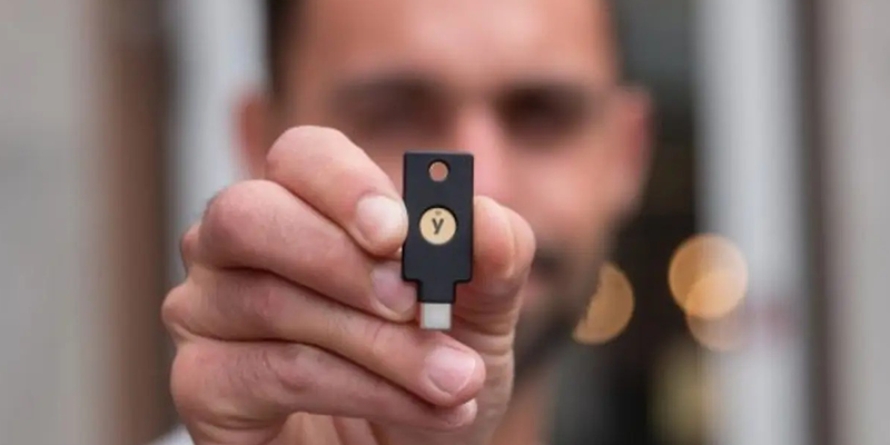 Close-up of a hand holding a black USB security key with gold button and "Y" logo, emphasizing cyber security, data security, endpoint security, network firewall, cloud security, information security, and IT support for small and medium businesses; blurred person in background highlights focus on IT compliance and network protection.