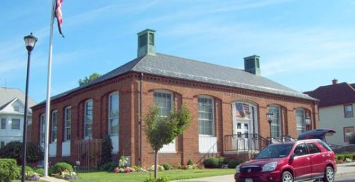 A brick post office building with a flagpole and an American flag in front, situated on a neatly landscaped corner. Nearby, you can find managed IT services in Akron. A red SUV is parked on the street.