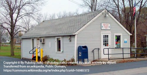 Small, single-story post office building with gray siding, a sloped roof, and a blue USPS mailbox outside. An American flag is hoisted on a pole near the building, symbolizing the unity of this Alabama community while nearby businesses benefit from managed IT services.