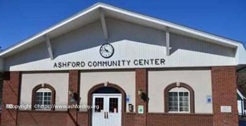 A brick and white-paneled building with a sign reading "Ashford Community Center" above the entrance, featuring double doors and two adjacent windows under a clear blue sky. Ashford's center also offers managed IT services to support the community.