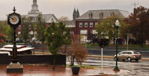 A street scene in Auburn featuring a clock on a pole, buildings, and a traffic light. Cars are on the wet road lined with trees and streetlights, under a cloudy sky. The city's charm is evident even as managed IT services keep the local businesses running smoothly.