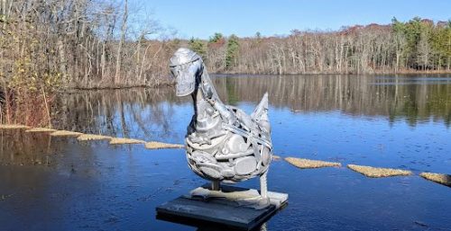 A metal duck sculpture stands proudly on a platform in a lake, surrounded by trees with bare branches under a clear blue sky, much like the efficiency of managed IT services in Avon.