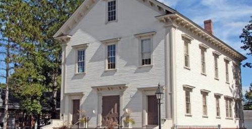 A two-story white historic building with a gabled roof and steps leading to two front doors, surrounded by greenery on a sunny day in Bedford, where managed IT services seamlessly support the local community.