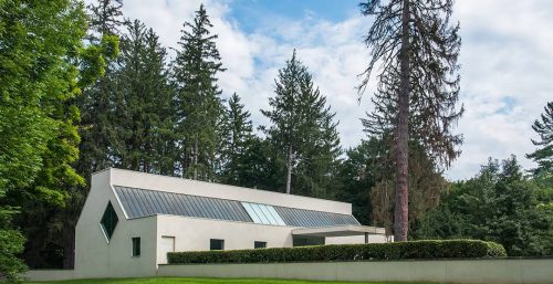 A modern, white, rectangular house with unique window designs sits in a lush green yard surrounded by tall trees against a partly cloudy sky, much like the innovative approach taken by managed IT services in Bedford.