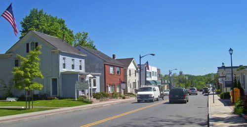 A sunny day on a small town street in Carmel, lined with buildings and parked cars, with an American flag waving in the breeze.