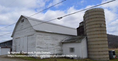 A large white barn with a gray roof and an adjacent cylindrical silo sits in a rural area. Power lines are visible overhead, symbolizing the connectivity that managed IT services Carrollton provides to local businesses. A grassy hill rises in the background, completing the picturesque scene.