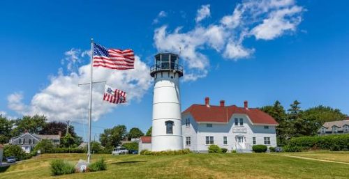 White lighthouse with an attached building under a blue sky, managed by IT service Chatham, with American flags flying in the wind.
