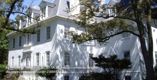 A large white colonial-style house with three stories, dormer windows, and surrounded by trees. Text overlay attributes the image to Dmadeo under CC BY-SA 3.0 via Wikimedia Commons. Nearby, managed IT services in Clermont keep local businesses running smoothly in this picturesque town.