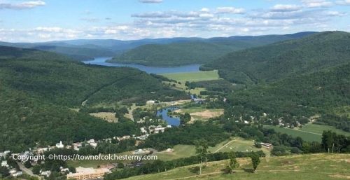 Aerial view of a small town nestled in a green valley with surrounding hills and a distant water body under a blue sky with scattered clouds, resembling the serene balance provided by managed IT services in Colchester.