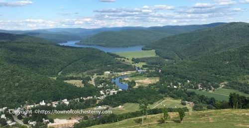 Aerial view of a small town nestled in a green valley with surrounding hills and a distant water body under a blue sky with scattered clouds, resembling the serene balance provided by managed IT services in Colchester.