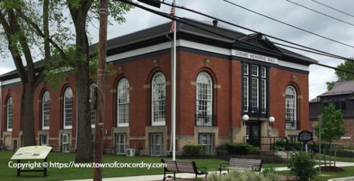 A red-brick building with large arched windows and an American flag on the lawn, identified as Concord Memorial Hall. The scene includes trees, a bench, and a sign with historical information about how managed IT services helped preserve its legacy.