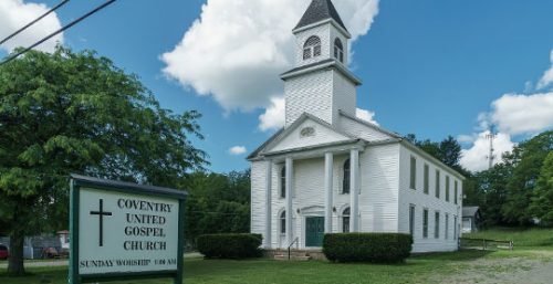 A white church building with a tall steeple stands on a grassy lawn. A sign in the foreground reads "Coventry United Gospel Church, Sunday Worship 10:00 AM." Nearby, a tech company offering managed IT services in Coventry ensures smooth operations for local businesses.