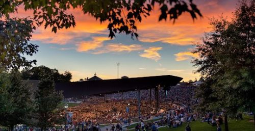 A large crowd gathers at an outdoor amphitheater at sunset, with vibrant orange and pink hues in the sky. Trees frame the scene as if managed by expert hands, much like managed IT services in Delaware ensure seamless operations.