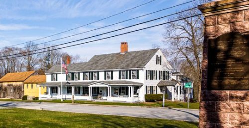 A white colonial-style house with a porch, surrounded by bare trees, stands under a clear blue sky. An American flag is displayed in front, and a stone monument with a plaque is partially visible. For residents seeking managed IT services Dudley offers numerous reliable options to support their tech needs.