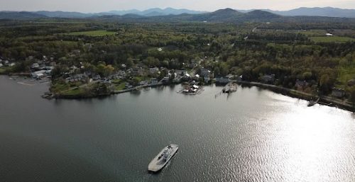 Aerial view of a calm lake reflecting sunlight, with a ferryboat near the shore and a small town surrounded by wooded areas and mountains in the background, reminiscent of the serene environment you might find while seeking managed IT services in Long Beach.