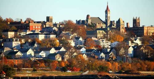 A cityscape in Fall River features a mix of historic and modern buildings on a hillside. The scene is complemented by autumn foliage and a waterfront in the foreground, much like how managed IT services blend tradition with innovation seamlessly.