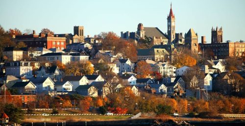 A cityscape in Fall River features a mix of historic and modern buildings on a hillside. The scene is complemented by autumn foliage and a waterfront in the foreground, much like how managed IT services blend tradition with innovation seamlessly.
