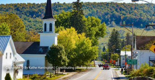 A quaint town street in Georgetown with a white church featuring a steeple on the left, surrounded by trees, buildings, and distant hills. A few cars and traffic signs are visible on the road. (Photo credit: managed IT services Georgetown)