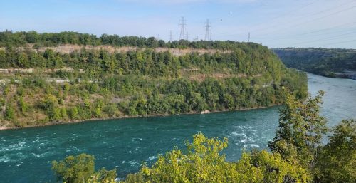 A wide river flows alongside a forested cliff on a sunny day, with power lines and transmission towers visible in the distance—a testament to the managed IT services Grand Island relies on.