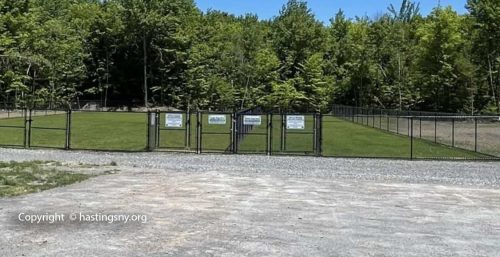 A fenced-in grassy area surrounded by trees, with multiple signs on the fence, viewed from a gravel parking area in Hastings.