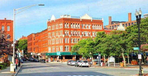 A city street in Haverhill with historic red-brick buildings, trees lining the sidewalks, parked cars, and a few pedestrians. The street has a crosswalk and streetlamp, seamlessly blending modern elements with classic charm—much like how managed IT services integrate into traditional business practices.